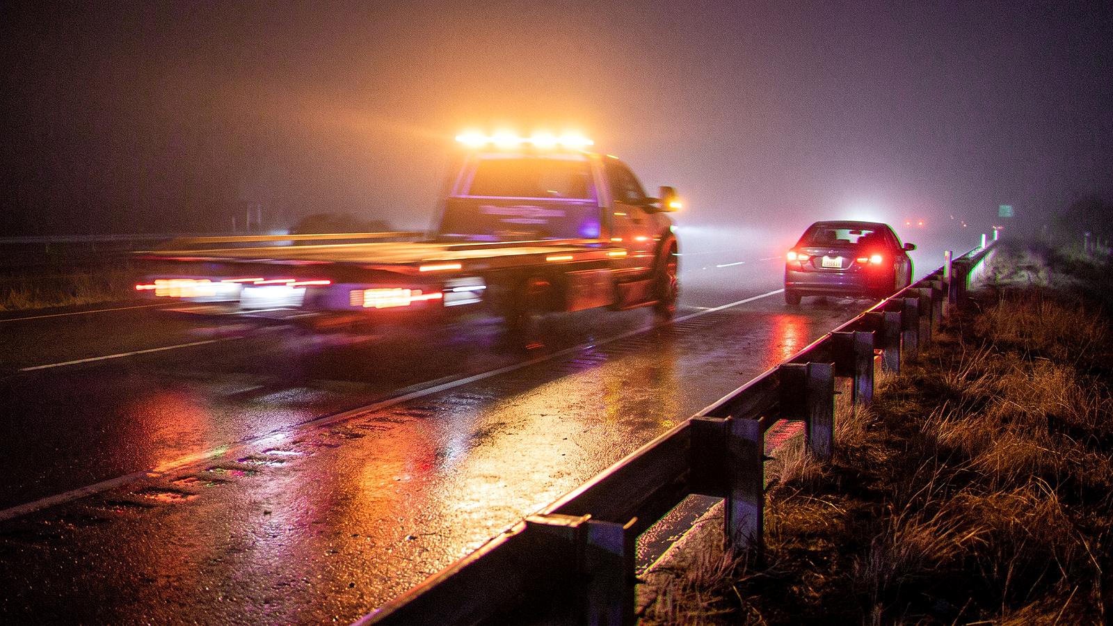 A tow truck's amber light bar on a fog-wet freeway shoulder at night, a stopped car's tail lights ahead